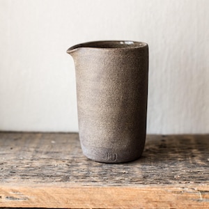 May include: A handcrafted, speckled brown ceramic pitcher with a pouring spout. The cylindrical vessel has a rounded base and a small, stamped maker's mark. It sits on a rustic wooden surface, showcasing its earthy tones and minimalist design.