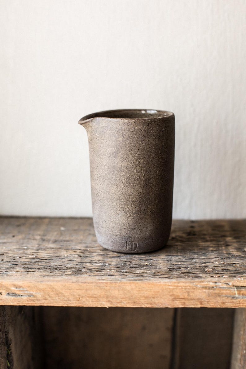 May include: A handcrafted, speckled brown ceramic pitcher with a pouring spout. The cylindrical vessel has a rounded base and a small, stamped maker's mark. It sits on a rustic wooden surface, showcasing its earthy tones and minimalist design.