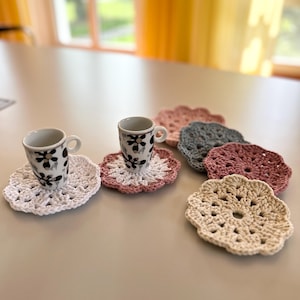 Può includere: Due tazze da caffè in ceramica bianca con motivi floreali neri si trovano su sottobicchieri all'uncinetto. I sottobicchieri sono bianchi, rosa e beige.