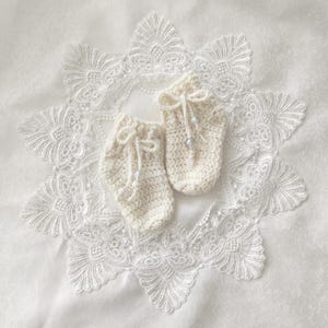 Może przedstawiać: Para kremowych, szydełkowych bucików dla niemowląt ze sznurkowymi zamknięciami i małymi akcentami z koralików. Buciki są prezentowane na białej, okrągłej serwetce koronkowej ze skomplikowanymi wzorami kwiatowymi, tworząc delikatną, ręcznie robioną estetykę.