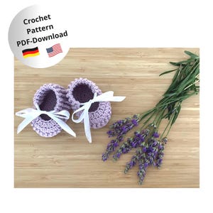 Könnte beinhalten: Ein Paar lila gehäkelte Babyschuhe mit weißen Schleifenbändern. Das Bild zeigt einen Lavendelzweig und eine kreisförmige Grafik mit dem Text "Crochet Pattern PDF-Download" und den Flaggen Deutschlands und der Vereinigten Staaten.