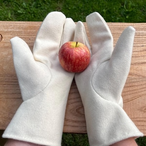 Op de afbeelding: Een paar off-white, effen handschoenen met een gladde, viltachtige textuur, samengehouden in een komvormige houding. Tussen de handpalmen van de handschoenen rust een enkele, ronde, rood-gele appel met een zichtbaar steeltje bovenaan.