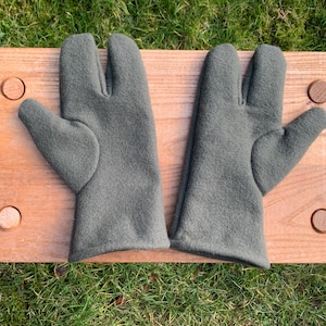 May include: Two gray oven mitts with a thumb and two finger slots, resting on a wooden cutting board with four wooden pegs. The mitts are made of a soft, textured material.