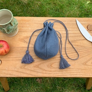 May include: A blue drawstring pouch with tassels, a mug with a face design, a red apple, and a knife with a wooden handle are on a wooden table. The table is outdoors on a grassy surface.