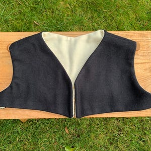 Pode incluir: Um colete reversível preto e creme com design de decote em V. O colete é feito de um tecido macio e tem um estilo simples e clássico. O colete é exibido em uma superfície de madeira contra um fundo de grama verde.