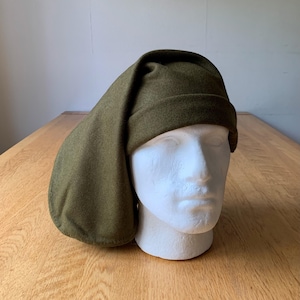 Può includere: Cappello medievale verde oliva con una lunga falda a punta. Il cappello è realizzato in un materiale morbido simile al feltro ed è esposto su una testa di manichino bianca. Il design del cappello ricorda i copricapi storici.