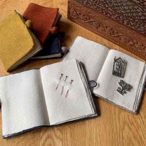 May include: A collection of small, colorful felt books with white pages, some open to display pins and decorative metal charms. The books are in shades of yellow, rust, and blue. A carved wooden box is in the background.
