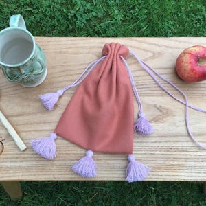May include: A peach-colored drawstring bag with lavender tassels and drawstrings. A wooden spoon, a green ceramic mug, and a red apple are arranged on a wooden surface, creating a rustic aesthetic.