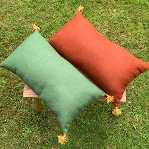 May include: Two rectangular cushions, one in a muted green and the other in a rust-orange hue, rest on a small wooden stool. Each cushion has a tassel at each corner. The cushions are on a grassy surface.