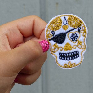Könnte beinhalten: Ein weißer gestickter Aufnäher mit einem goldenen und schwarzen Zucker Totenkopf mit einem schwarzen Augenklappen und einem Blumendesign.