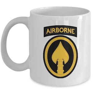 May include: White ceramic coffee mug with a gold and black airborne insignia. The insignia features a spearhead with a rope border and the word "AIRBORNE" in a black banner.