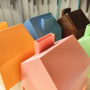 May include: A selection of brightly coloured, geometric house-shaped objects. The houses are painted in shades of orange, blue, pink, brown, and green. Each house has a rectangular chimney. The objects appear to be decorative or functional.