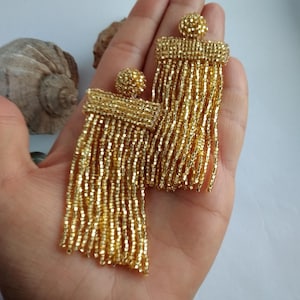 Könnte beinhalten: Ein Paar goldene Perlen-Quastenohrringe. Die Ohrringe sind lang und haben eine Fransen aus Perlen, die von einer rechteckigen Basis herabhängen.