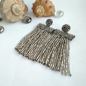 May include: A pair of silver beaded tassel earrings. The earrings have a small, round, beaded stud at the top and a rectangular beaded section from which the long tassels hang. The earrings are displayed on a white surface with seashells and glass beads.