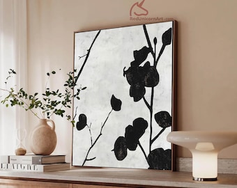 May include: A framed abstract painting with black floral silhouettes against a textured white background. The artwork is displayed in a room with a neutral color palette, complemented by a vase with greenery and a modern lamp.