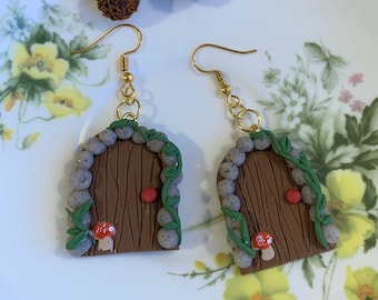 May include: A pair of brown and green fairy door earrings with gold hooks. The doors are made of brown clay with a red mushroom and green vines. The doors are surrounded by grey stones.