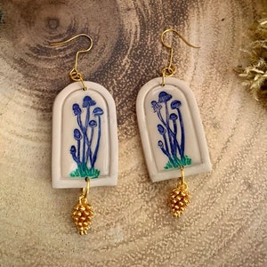 Könnte beinhalten: Ein Paar weiße Keramikohrringe mit einem blauen und grünen Pilzdesign und goldenen Akzenten. Die Ohrringe sind bogenförmig und haben einen kleinen goldenen Quasten, der an der Unterseite hängt.