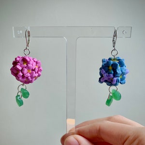 May include: Handmade earrings with floral designs. One is pink, the other blue, both with green leaf accents. The earrings are displayed on a clear stand.