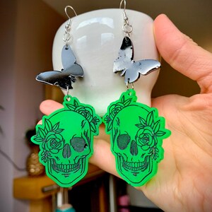 May include: A pair of green skull earrings with black butterfly charms. The skulls are decorated with black floral details.