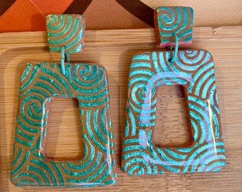 Puede incluir: Un par de pendientes llamativos con forma rectangular. Los pendientes están hechos de arcilla marrón con un patrón de remolino verde. Los pendientes tienen un acabado brillante y brillante.