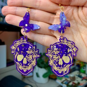 May include: Purple and gold glitter skull earrings with a butterfly charm. The skull is decorated with gold floral details.