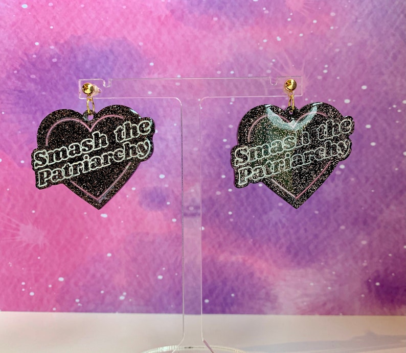 May include: A pair of heart-shaped earrings with the text "Smash the Patriarchy". The earrings are black with glitter and a pink outline. They hang from gold-colored posts.