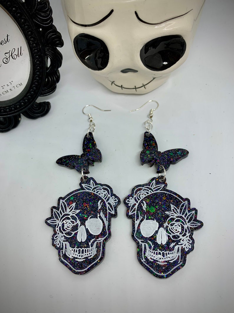 May include: Black glitter skull earrings with white floral details and black butterfly charms.