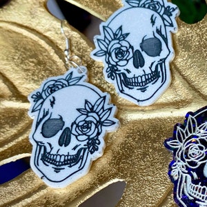 May include: A pair of white skull earrings with black floral details. The skulls have a glitter finish and are accented with black roses.