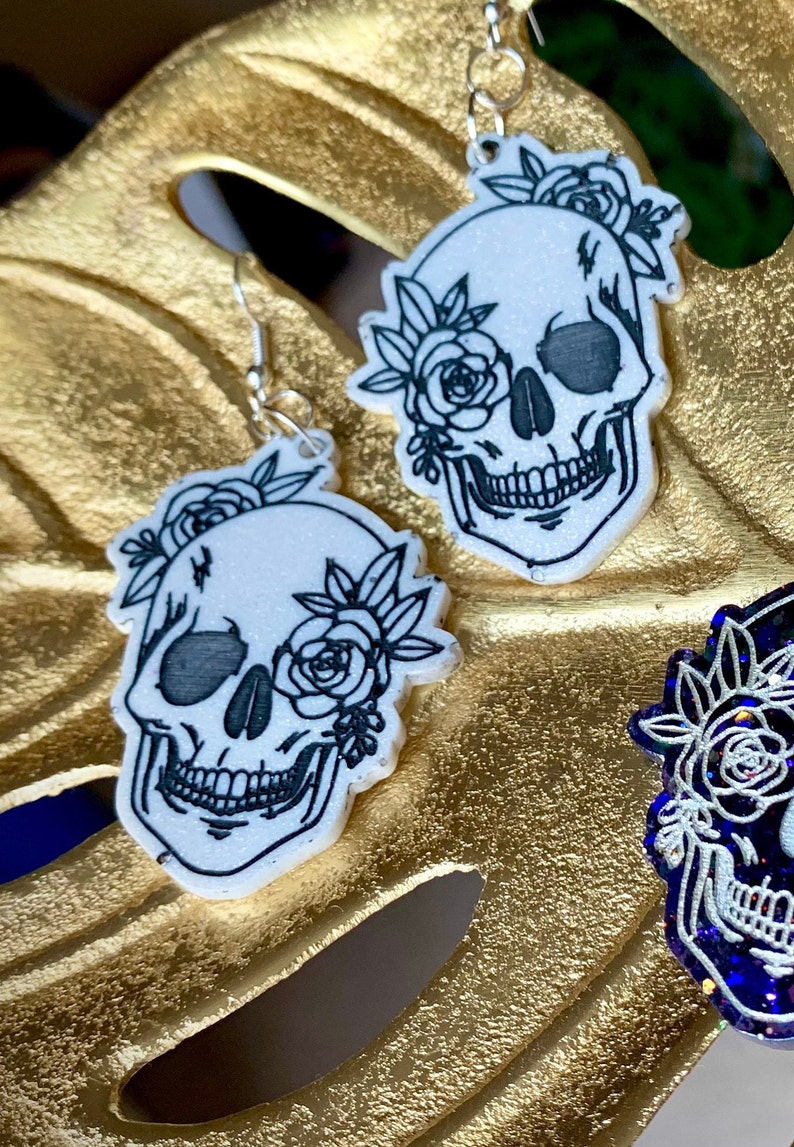 May include: A pair of white skull earrings with black floral details. The skulls have a glitter finish and are accented with black roses.