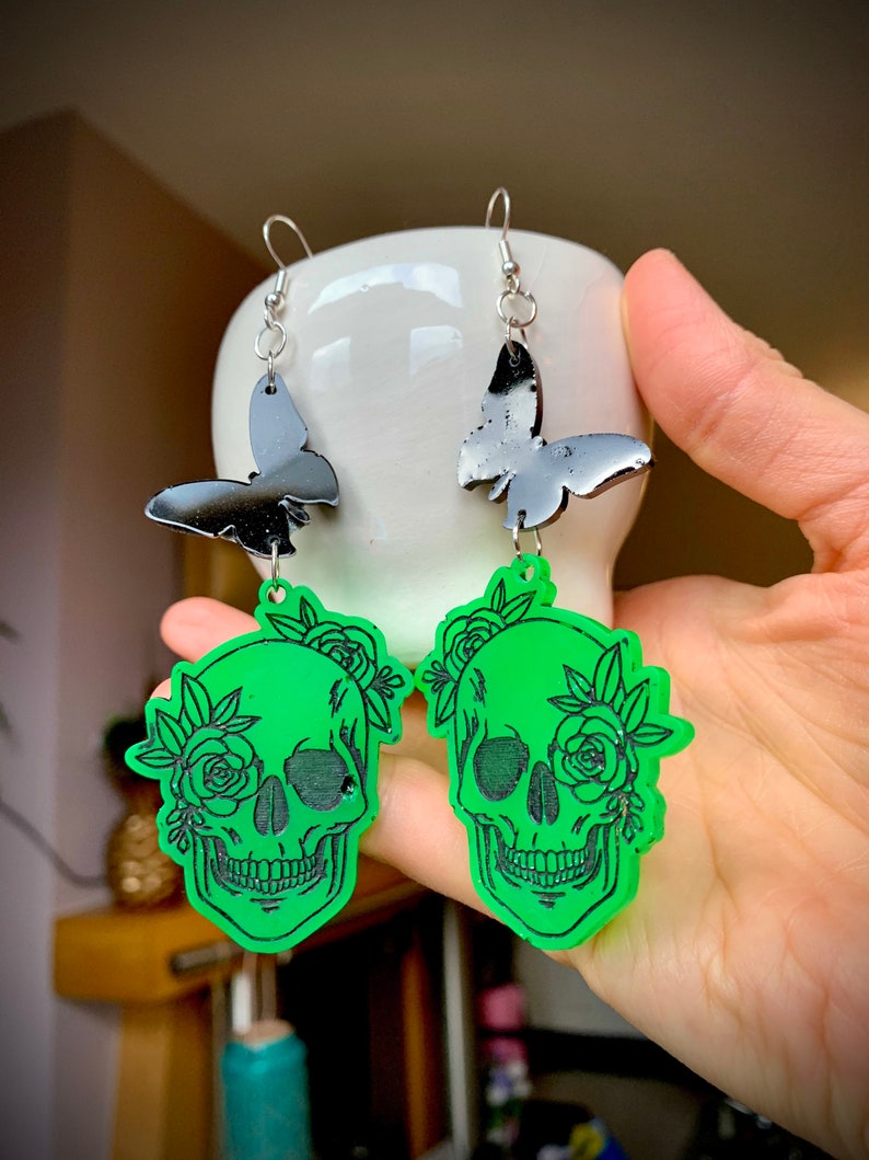 May include: A pair of green skull earrings with black butterfly charms. The skulls are decorated with black floral details.