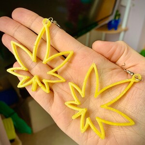 Könnte beinhalten: Ein Paar gelbe Ohrringe, die wie Marihuana-Blätter geformt sind. Die Ohrringe sind aus einem glatten, glänzenden Material gefertigt und haben ein schlichtes, modernes Design.