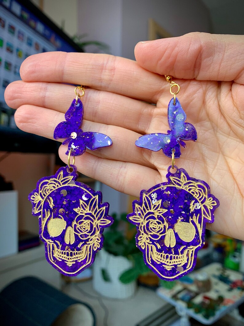 May include: Purple and gold glitter skull earrings with a butterfly charm. The skull is decorated with gold floral details.