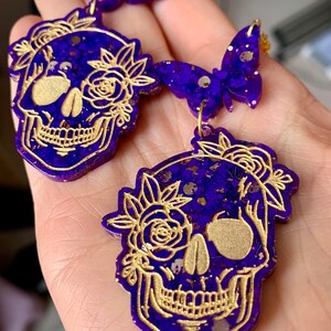 May include: Purple and gold glitter resin earrings featuring a skull design with floral accents. The skull is outlined in gold and has a detailed design. The earrings are dangle style with a butterfly charm.