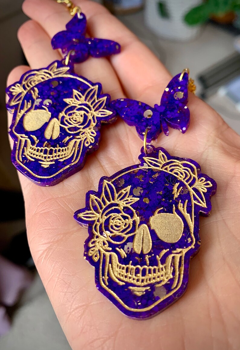 May include: Purple and gold glitter resin earrings featuring a skull design with floral accents. The skull is outlined in gold and has a detailed design. The earrings are dangle style with a butterfly charm.