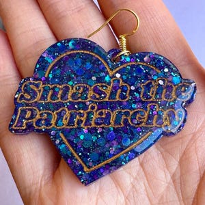 May include: A heart-shaped earring with a gold-colored hook. The earring is filled with blue and purple glitter and features the phrase "Smash the Patriarchy" in gold lettering.