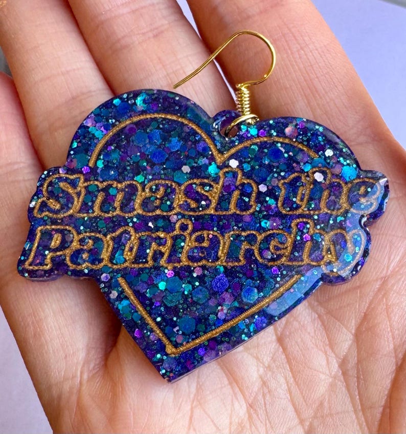 May include: A heart-shaped earring with a gold-colored hook. The earring is filled with blue and purple glitter and features the phrase "Smash the Patriarchy" in gold lettering.