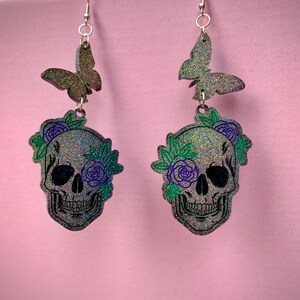 May include: A pair of silver dangle earrings featuring a black glitter skull with green and purple floral accents and a black glitter butterfly charm.