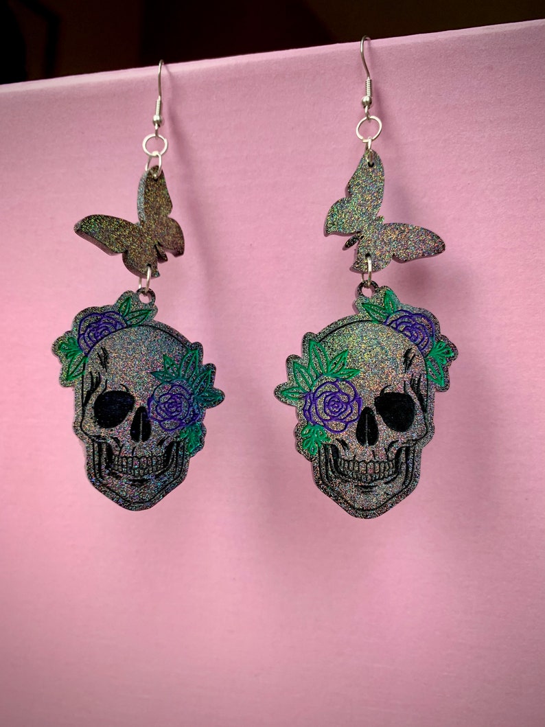 May include: A pair of silver dangle earrings featuring a black glitter skull with green and purple floral accents and a black glitter butterfly charm.
