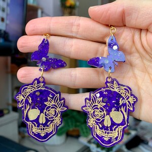 May include: Purple and gold glitter skull earrings with a butterfly charm. The skull is decorated with gold floral details.