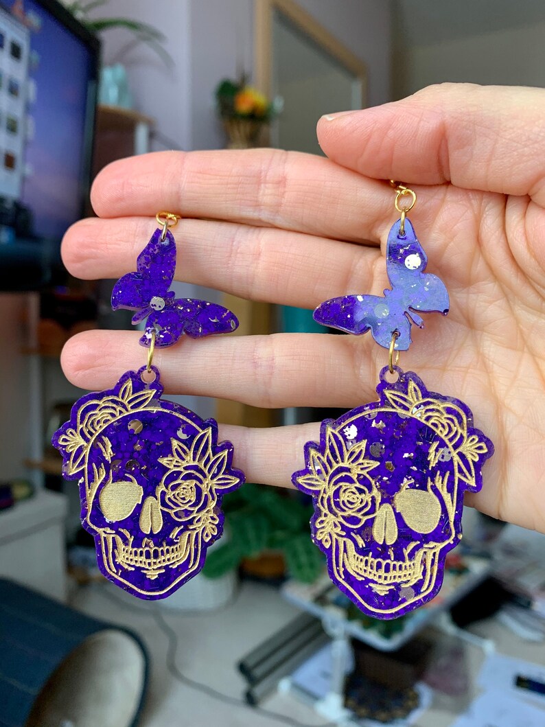 May include: Purple and gold glitter skull earrings with a butterfly charm. The skull is decorated with gold floral details.