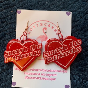 May include: A pair of red heart-shaped earrings with the text "Smash the Patriarchy" in white. The earrings have gold-colored hooks and are displayed on a white card with a flamingo logo and shop information.