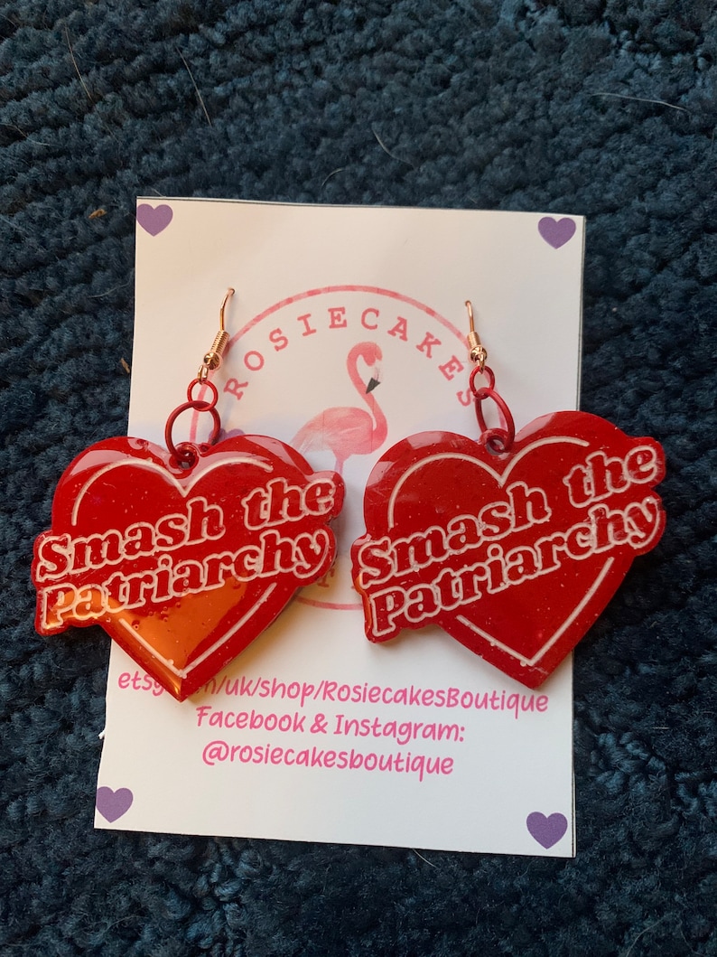 May include: A pair of red heart-shaped earrings with the text "Smash the Patriarchy" in white. The earrings have gold-colored hooks and are displayed on a white card with a flamingo logo and shop information.