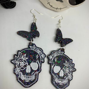 May include: A pair of black glitter skull earrings with white floral details and black glitter butterfly charms. The earrings have silver hooks.