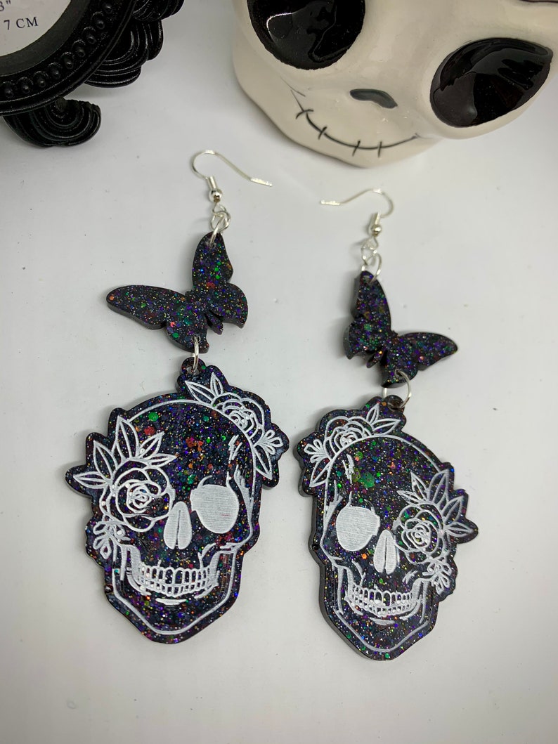 May include: A pair of black glitter skull earrings with white floral details and black glitter butterfly charms. The earrings have silver hooks.