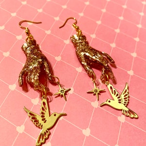 Puede incluir: Pendientes de mano de purpurina dorada con un colibrí dorado y un encanto de estrella que cuelgan de cada mano.