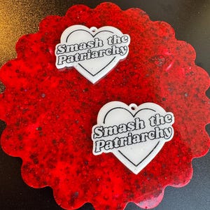 May include: Two white heart-shaped pendants with the text "Smash the Patriarchy" on a red, scalloped coaster. The coaster has a speckled, glittery appearance. The pendants have a small hole at the top for hanging.