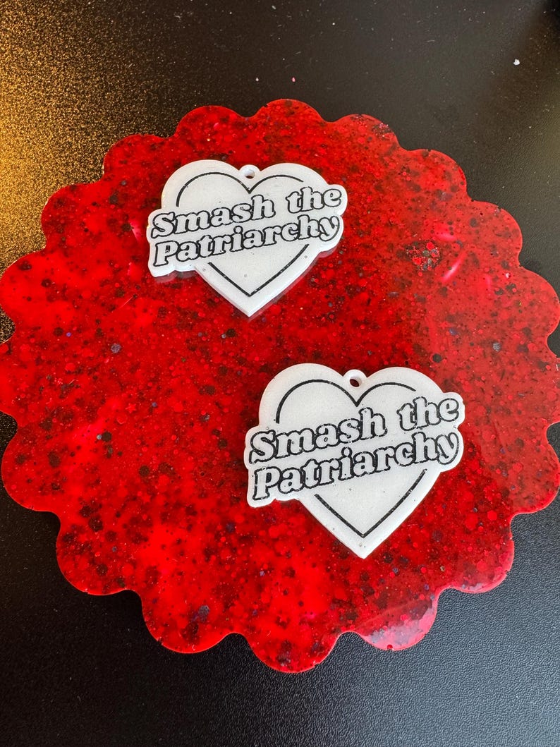 May include: Two white heart-shaped pendants with the text "Smash the Patriarchy" on a red, scalloped coaster. The coaster has a speckled, glittery appearance. The pendants have a small hole at the top for hanging.