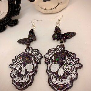 May include: A pair of black glitter skull earrings with white floral details and black glitter butterfly charms. The earrings have silver hooks.