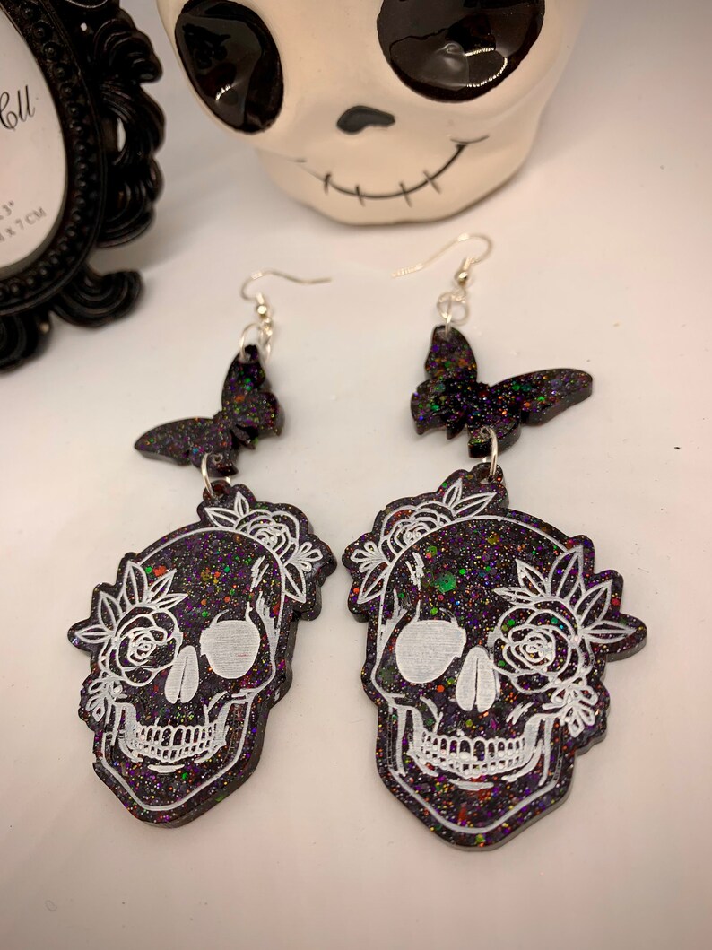 May include: A pair of black glitter skull earrings with white floral details and black glitter butterfly charms. The earrings have silver hooks.