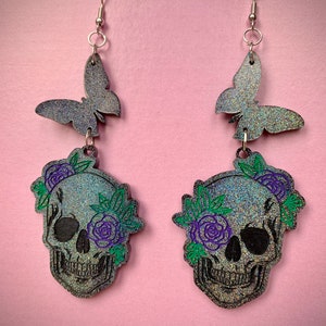 May include: A pair of silver dangle earrings with a black glitter skull with purple and green floral accents and a black glitter butterfly charm.